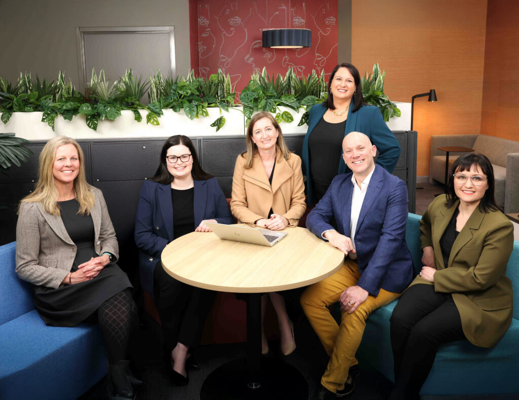 A group of six professionals — five women and one man — posed together in a modern office lounge. Five are seated around a round wooden table and one woman stands at the back. All are smiling and dressed in business attire.